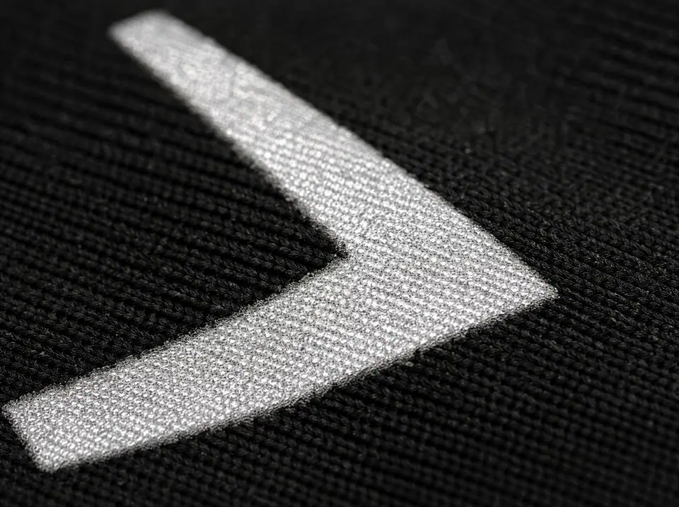 Macro of a retroreflective heat transfer on black technical fabric flaring silver-white under a direct beam, glass-bead structure visible
