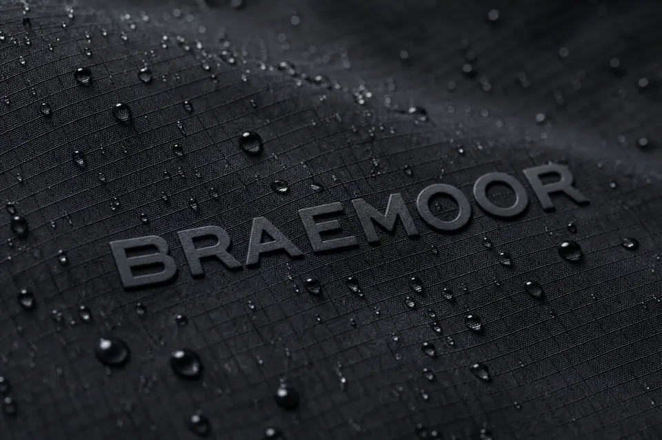 Macro of black coated ripstop shell fabric: a raised tonal silicone brand mark holding its edge while water beads across the water-repellent face